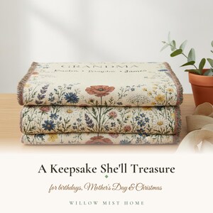 May include: A folded woven throw blanket featuring a cream-colored background with a colorful floral pattern of poppies, daisies, and lavender. The top section displays the text GRANDMA followed by the names Kristen, Brayden, and James.