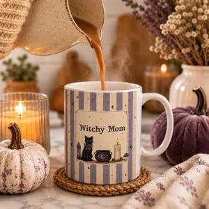 May include: A white ceramic mug with a handle, featuring vertical light purple stripes and a central rectangular graphic with the text "Witchy Mom". The illustration includes a black cat, a purple crystal, a cauldron, and a lit candle.