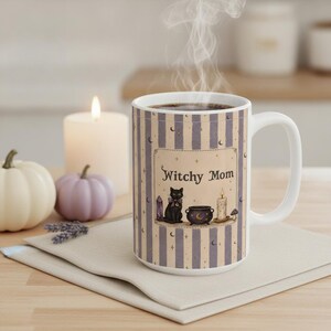 May include: A white ceramic mug featuring a vertical striped pattern in beige and muted purple. The center displays a rectangular graphic with the text Witchy Mom, a black cat, a purple crystal, a small cauldron, and a lit candle with stars and moons.