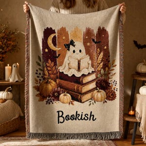 May include: A woven throw blanket featuring a ghost with a black bow reading a book while sitting on a stack of three books. The design includes pumpkins, autumn leaves, a crescent moon, and bats. The word Bookish is printed at the bottom.