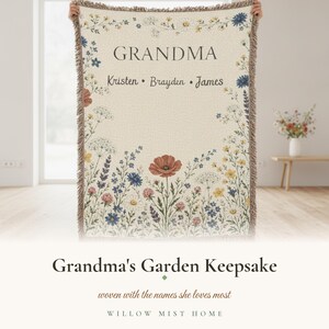 May include: A cream-colored woven throw blanket featuring a floral border of wildflowers in shades of red, blue, and yellow. The text GRANDMA is printed at the top, followed by the names Kristen, Brayden, and James in a dark serif font.