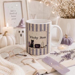 May include: A white ceramic mug featuring a vertical lavender and cream striped pattern. The center design displays a black cat, a purple cauldron, a candle, and the text Witchy Mom. The mug is decorated with small stars and crescent moon icons.