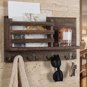 May include: A dark brown wooden wall organizer with a shelf, mail slots, and hooks. The shelf holds a glass with pink crystals and the hooks hold keys, sunglasses, and a scarf. The mail slots contain cards and papers.