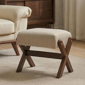 May include: A beige upholstered ottoman with a rectangular top and dark brown wooden X-shaped legs. The ottoman is placed on a light-colored rug, and a cream-colored armchair is visible in the background.