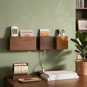 Può includere: Tre organizer da parete in legno in varie tonalità di marrone. Contengono un telecomando, una carta con il testo "Limited Romance", un telefono con cavo di ricarica, penne e occhiali. Sono visibili anche un calendario da scrivania e dei libri.