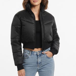 May include: A black, cropped puffer jacket with a high collar and ribbed cuffs. The jacket features a chevron design on the front and a ribbed hem. The model is wearing the jacket with a black top and blue jeans.