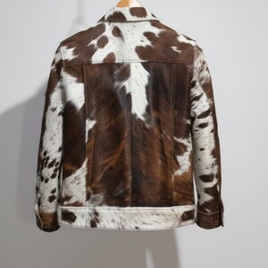 Puede incluir: Chaqueta de piel de vaca marr&oacute;n y blanca con botones y cuello. La chaqueta tiene un dise&ntilde;o cl&aacute;sico con un patr&oacute;n natural &uacute;nico. La parte trasera de la chaqueta es visible, mostrando las variaciones de textura y color de la piel.