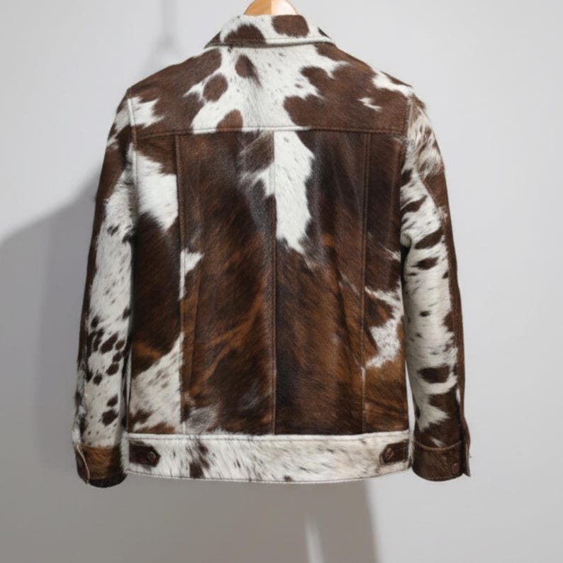 Puede incluir: Chaqueta de piel de vaca marr&oacute;n y blanca con botones y cuello. La chaqueta tiene un dise&ntilde;o cl&aacute;sico con un patr&oacute;n natural &uacute;nico. La parte trasera de la chaqueta es visible, mostrando las variaciones de textura y color de la piel.