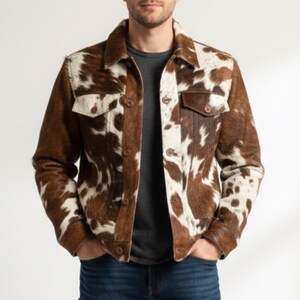 Puede incluir: Chaqueta de piel de vaca marr&oacute;n y blanca con cierre de botones, dos bolsillos en el pecho y cuello. La chaqueta tiene un estilo cl&aacute;sico de chaqueta vaquera con un patr&oacute;n manchado &uacute;nico. Las mangas son largas. Se lleva sobre una camiseta gris y vaqueros azules.