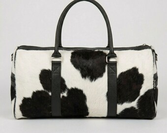 May include: A duffel bag featuring a black and white cow print pattern with a textured hair-on-hide finish. The bag includes two black leather handles, black leather trim, and silver-toned metal hardware attachments for the handles and side straps.
