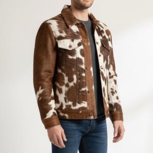 Puede incluir: Chaqueta de piel de vaca marr&oacute;n y blanca con cuello cl&aacute;sico, dos bolsillos en el pecho y cierre de botones. La chaqueta tiene mangas y ribetes marrones, y se usa con una camisa gris y vaqueros azules.