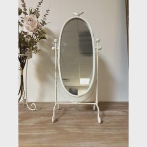 May include: An off-white, oval vanity mirror with a bird perched on top. The mirror is supported by a decorative metal frame with a stand. The mirror is approximately 15 inches tall and is a decorative home accent.