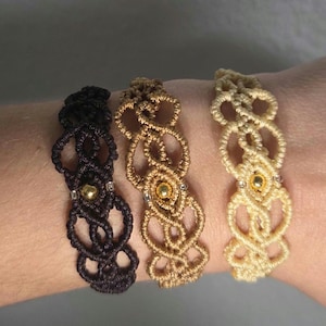 May include: Three macrame bracelets in black, gold, and cream colors. Each bracelet features an intricate woven design with a central gold bead and small silver beads. The bracelets are displayed on a wrist, showcasing the handcrafted details.