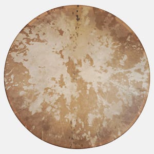 May include: A circular drumhead with a natural, mottled appearance. The surface is mainly tan with irregular patches of lighter, off-white colouration. The drum's edge is a darker brown, and the overall texture suggests a rawhide material.
