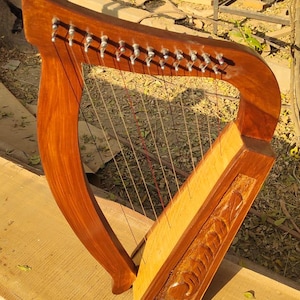 May include: A wooden harp with a warm brown finish. The instrument features a curved neck and a soundbox with carved details. The strings are visible, and the harp is resting on a wooden surface.