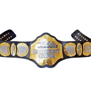 Könnte beinhalten: Ein TNA Wrestling Heavyweight Champion Gürtel. Der Gürtel hat eine goldfarbene Mittelplatte mit den Worten "TNA Wrestling Heavyweight Champion" und goldfarbene Seitenteile. Der Gürtel ist schwarz mit goldenen Akzenten.