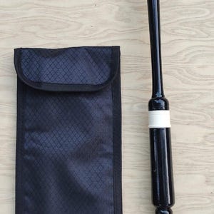 Puede incluir: Una caña de gaita negra y una funda de transporte negra. La caña es negra con detalles blancos y una boquilla blanca. La funda de transporte es negra con un patrón de diamantes.