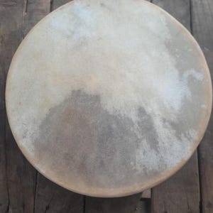 May include: A round, light-colored drum with a natural hide surface. The drumhead has a mottled appearance with variations in tone, ranging from off-white to light brown. The wooden frame is visible around the edge. The drum is likely used for music or ceremonial purposes.