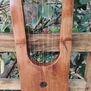 May include: A handcrafted wooden lyre with a warm brown finish. The instrument features a rounded top, a central sound hole, and multiple strings. It is set against a natural background.
