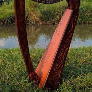 May include: A handcrafted wooden harp with detailed carvings, positioned in a grassy area beside a body of water. The dark brown wood of the harp contrasts with the light strings and the surrounding greenery. The harp is a musical instrument.