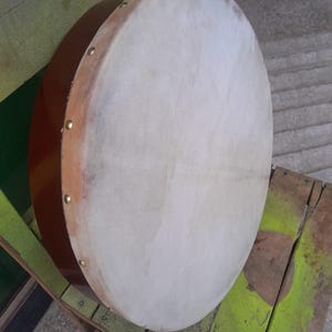 May include: A round, brown drum with a light-colored drumhead. The drum's wooden frame is visible, and it is secured with small, gold-colored tacks. The drum is resting on a green and brown wooden surface.