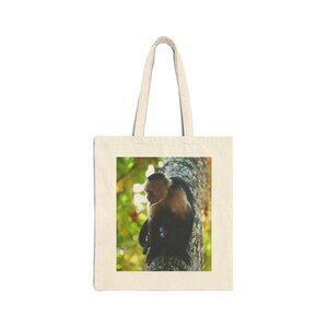 Peut inclure: Un sac fourre-tout en toile couleur crème avec une impression photographique rectangulaire sur le devant. L'image montre un singe capucin à tête blanche avec une fourrure noire sur le corps et les membres, perché sur un tronc d'arbre texturé.
