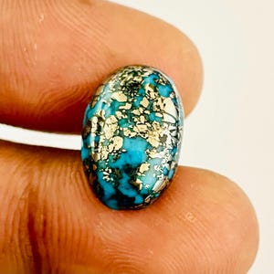 May include: Oval-shaped turquoise gemstone with a matrix of gold-colored pyrite inclusions. The stone is held between fingers, showcasing its polished surface and vibrant color. The gemstone's unique pattern and color make it a striking piece.