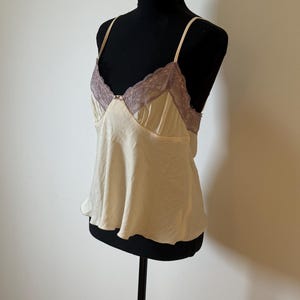 May include: Cream-colored camisole with thin straps and a V-neckline. The neckline is trimmed with lavender lace. The top is displayed on a black mannequin.