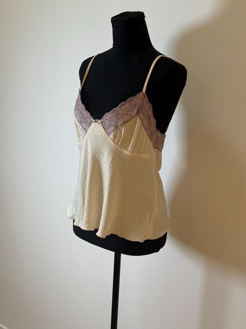 May include: Cream-colored camisole with thin straps and a V-neckline. The neckline is trimmed with lavender lace. The top is displayed on a black mannequin.