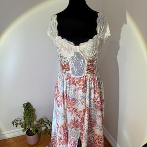 May include: A vintage-style dress on a black mannequin. The bodice features a white lace overlay and a sweetheart neckline. The skirt is a flowing, floral print in shades of pink, yellow, and blue on a white background. The dress has a high waist and a long, split skirt.