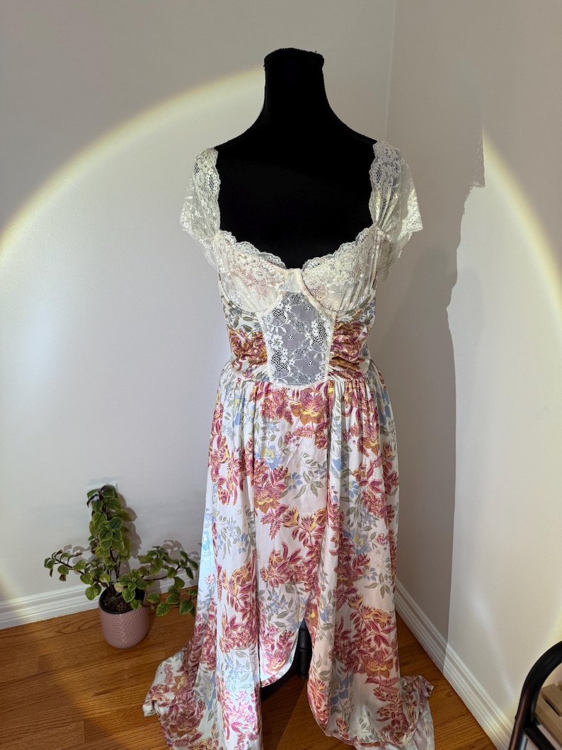 May include: A vintage-style dress on a black mannequin. The bodice features a white lace overlay and a sweetheart neckline. The skirt is a flowing, floral print in shades of pink, yellow, and blue on a white background. The dress has a high waist and a long, split skirt.