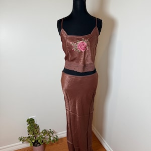 Puede incluir: Conjunto de dos piezas de sat&eacute;n marr&oacute;n sobre un maniqu&iacute; negro. La parte superior es una camiseta sin mangas con bordado de rosas y ribete de encaje. La falda a juego es larga hasta el suelo. El conjunto se exhibe sobre un suelo de madera contra una pared blanca.