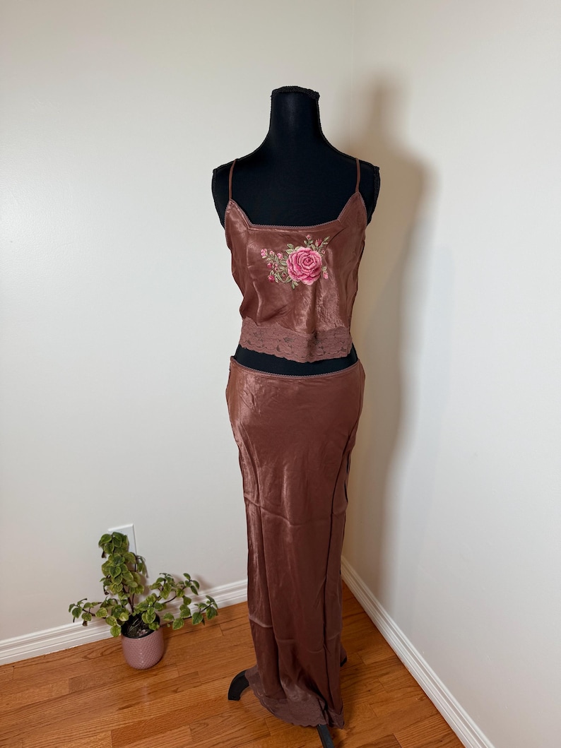 Puede incluir: Conjunto de dos piezas de sat&eacute;n marr&oacute;n sobre un maniqu&iacute; negro. La parte superior es una camiseta sin mangas con bordado de rosas y ribete de encaje. La falda a juego es larga hasta el suelo. El conjunto se exhibe sobre un suelo de madera contra una pared blanca.
