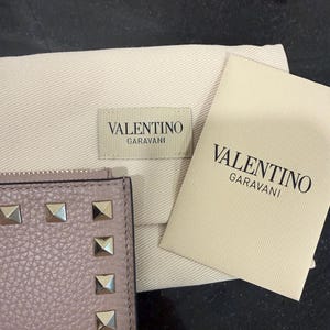 May include: A light purple leather wallet with silver-colored square studs. The wallet is next to a beige dust bag and a card with the text "VALENTINO GARAVANI".