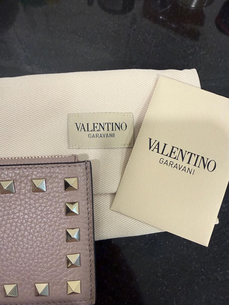 May include: A light purple leather wallet with silver-colored square studs. The wallet is next to a beige dust bag and a card with the text "VALENTINO GARAVANI".