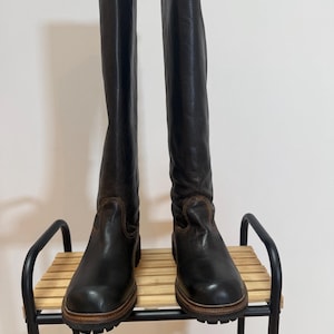 May include: A pair of knee-high black leather boots featuring a rounded toe, a thick brown welt, and a black lug sole. The boots have visible brown stitching around the vamp and a smooth, dark leather finish extending up to the calf.
