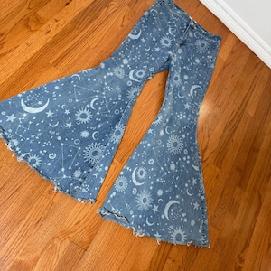 May include: Light blue denim bell-bottom jeans featuring a celestial pattern of moons, stars, and suns. The jeans have a distressed hem and a high waist. The flared legs create a retro, bohemian style.