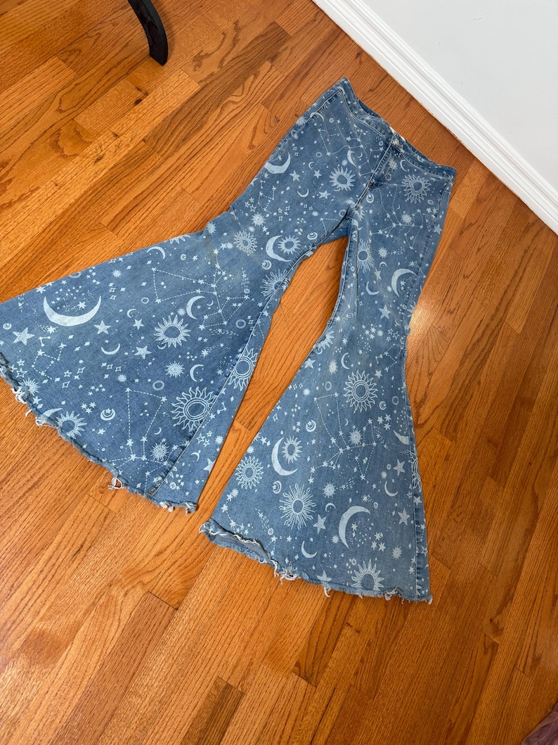 May include: Light blue denim bell-bottom jeans featuring a celestial pattern of moons, stars, and suns. The jeans have a distressed hem and a high waist. The flared legs create a retro, bohemian style.