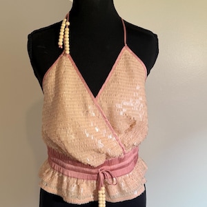 May include: A light pink sequined halter top with a wrap-style front and a gathered waist. The top features a pink satin trim and a beaded tie at the waist. The halter straps are also adorned with beads. The top is displayed on a black mannequin.