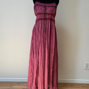 May include: A long, magenta dress displayed on a black mannequin. The dress has a gathered bodice with black trim and spaghetti straps. The skirt has a textured, pleated appearance, creating a soft, flowing silhouette. The dress is suitable for a variety of occasions.