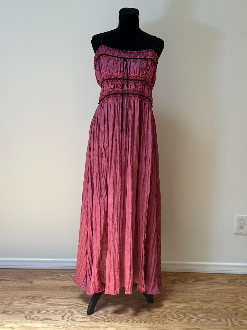 May include: A long, magenta dress displayed on a black mannequin. The dress has a gathered bodice with black trim and spaghetti straps. The skirt has a textured, pleated appearance, creating a soft, flowing silhouette. The dress is suitable for a variety of occasions.