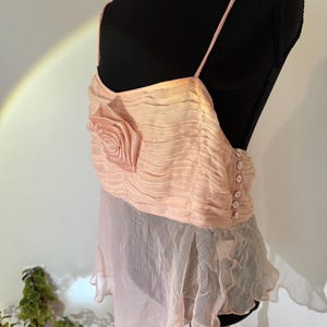 May include: A light peach-colored top with spaghetti straps and a rose-shaped embellishment. The top features a ruched bodice and a flowing, sheer skirt. The back has a button closure. The garment is displayed on a black mannequin.