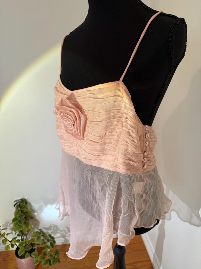 May include: A light peach-colored top with spaghetti straps and a rose-shaped embellishment. The top features a ruched bodice and a flowing, sheer skirt. The back has a button closure. The garment is displayed on a black mannequin.