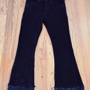 Puede incluir: Vaqueros negros de mezclilla con cintura alta y perneras acampanadas. Los vaqueros tienen un detalle de dobladillo deshilachado en la parte inferior de las perneras. Los vaqueros se muestran sobre una superficie de madera.