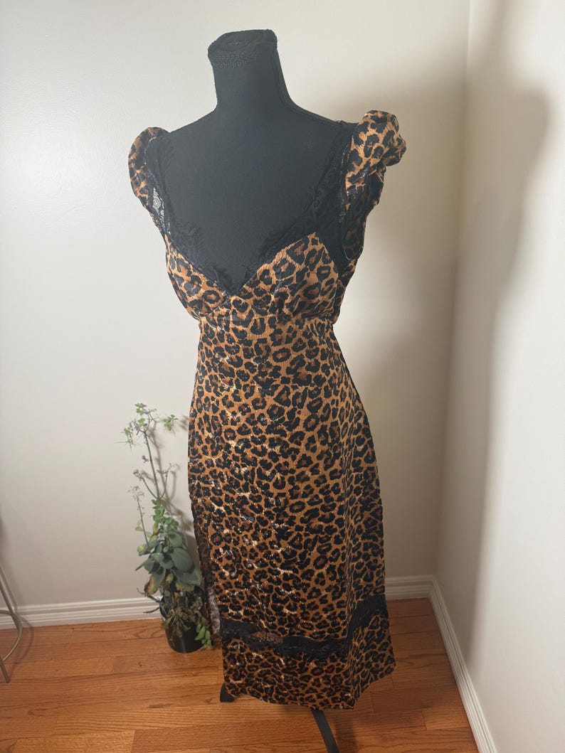 Puede incluir: Un vestido con estampado de leopardo con adornos y detalles de encaje negro. El vestido se exhibe en un maniqu&iacute; negro. El vestido tiene un escote en V y mangas cortas. El vestido llega a la rodilla.