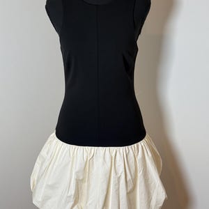 May include: A sleeveless black dress with a cream-colored, gathered bubble skirt. The dress is displayed on a black mannequin against a neutral background. The dress has a simple, elegant design.