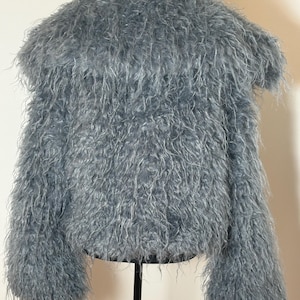May include: A light blue, fuzzy jacket with long sleeves. The jacket is made of a soft, shaggy material and has a round neckline. The jacket is displayed on a black stand.