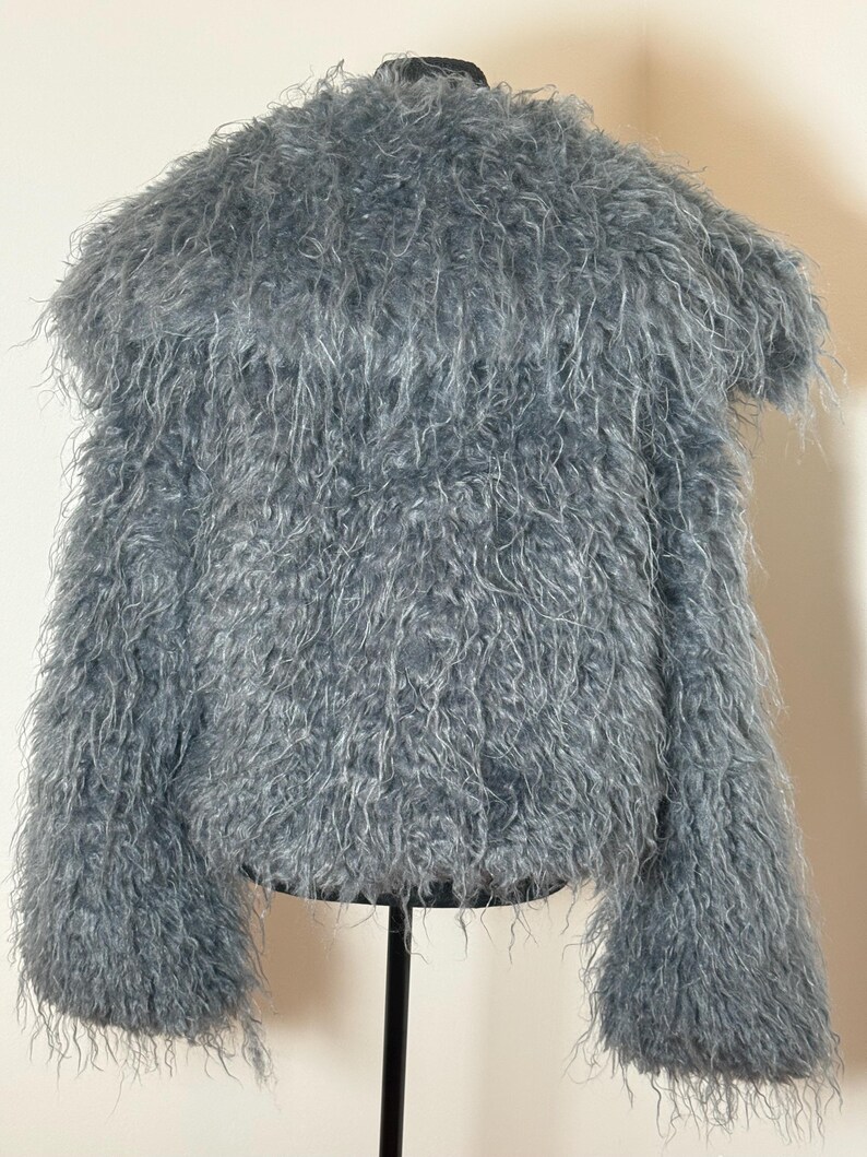 May include: A light blue, fuzzy jacket with long sleeves. The jacket is made of a soft, shaggy material and has a round neckline. The jacket is displayed on a black stand.