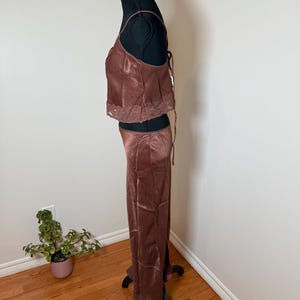 Puede incluir: Un conjunto de dos piezas de sat&eacute;n marr&oacute;n sobre un maniqu&iacute; negro. La parte superior es un camisola con tirantes finos y ribete de encaje. Los pantalones a juego son de pernera ancha. El conjunto se exhibe en un maniqu&iacute; negro contra una pared blanca.
