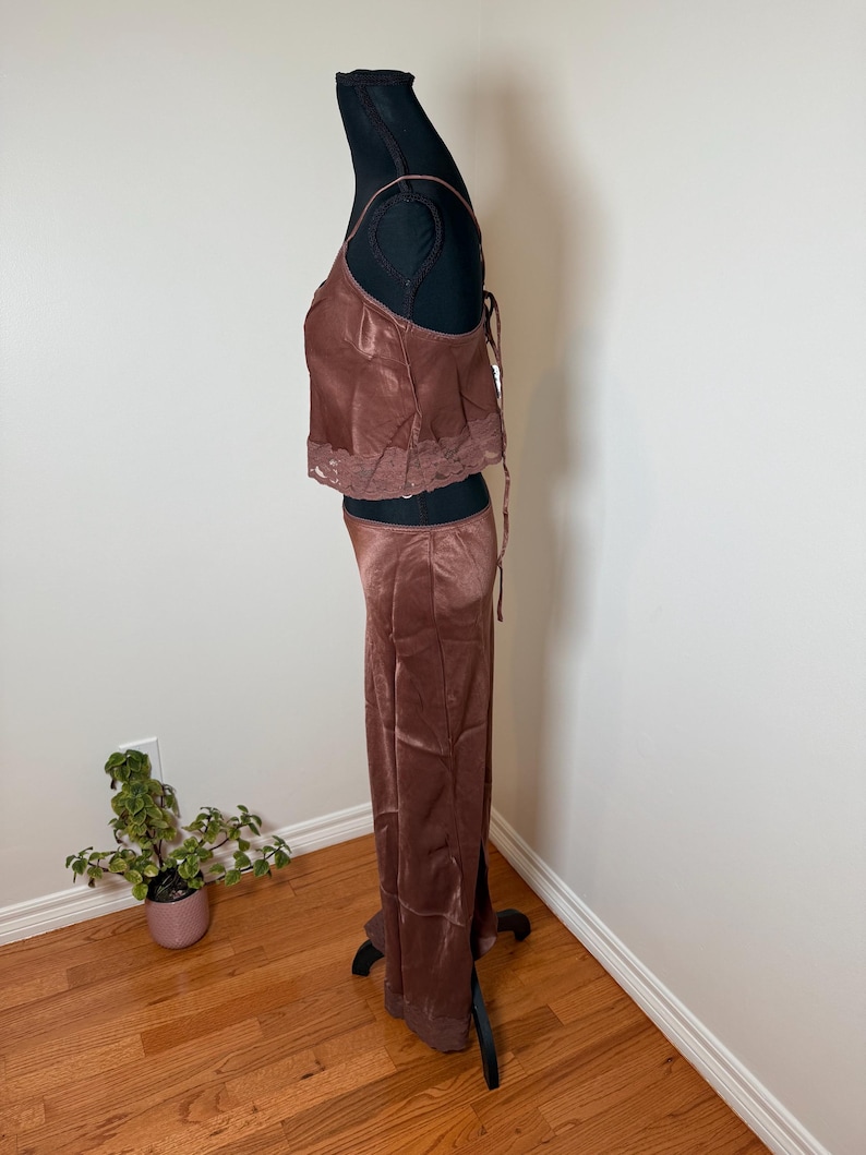 Puede incluir: Un conjunto de dos piezas de sat&eacute;n marr&oacute;n sobre un maniqu&iacute; negro. La parte superior es un camisola con tirantes finos y ribete de encaje. Los pantalones a juego son de pernera ancha. El conjunto se exhibe en un maniqu&iacute; negro contra una pared blanca.